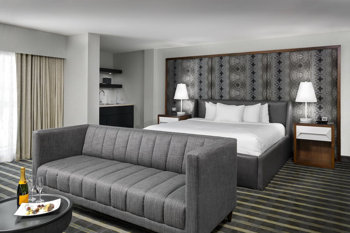 A modern hotel room with a grey sofa, a large bed, a nightstand with a lamp, and a small table with a bottle and glasses.