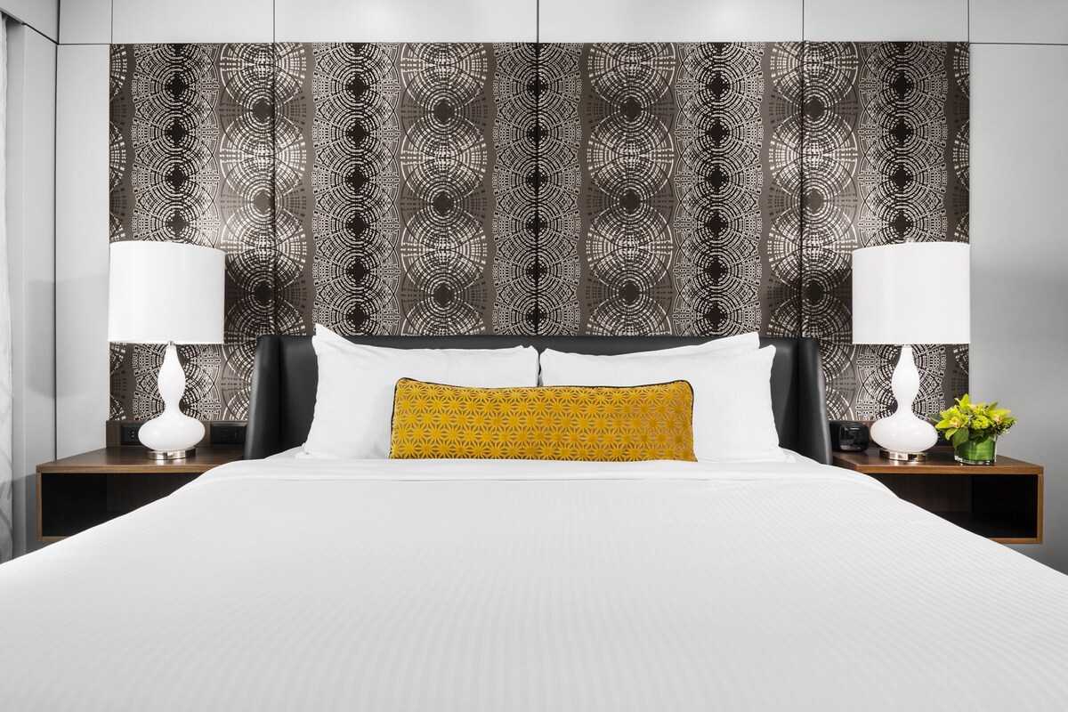 A hotel room with a large bed, two bedside tables with lamps, a patterned headboard, and a small vase with flowers.