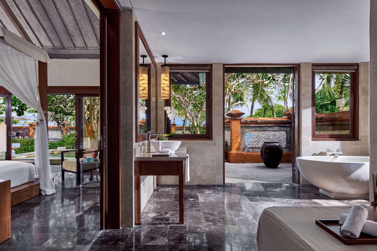 A modern bathroom with a large bathtub, a sink, and a mirror. There is a window with a view of palm trees and a pool.