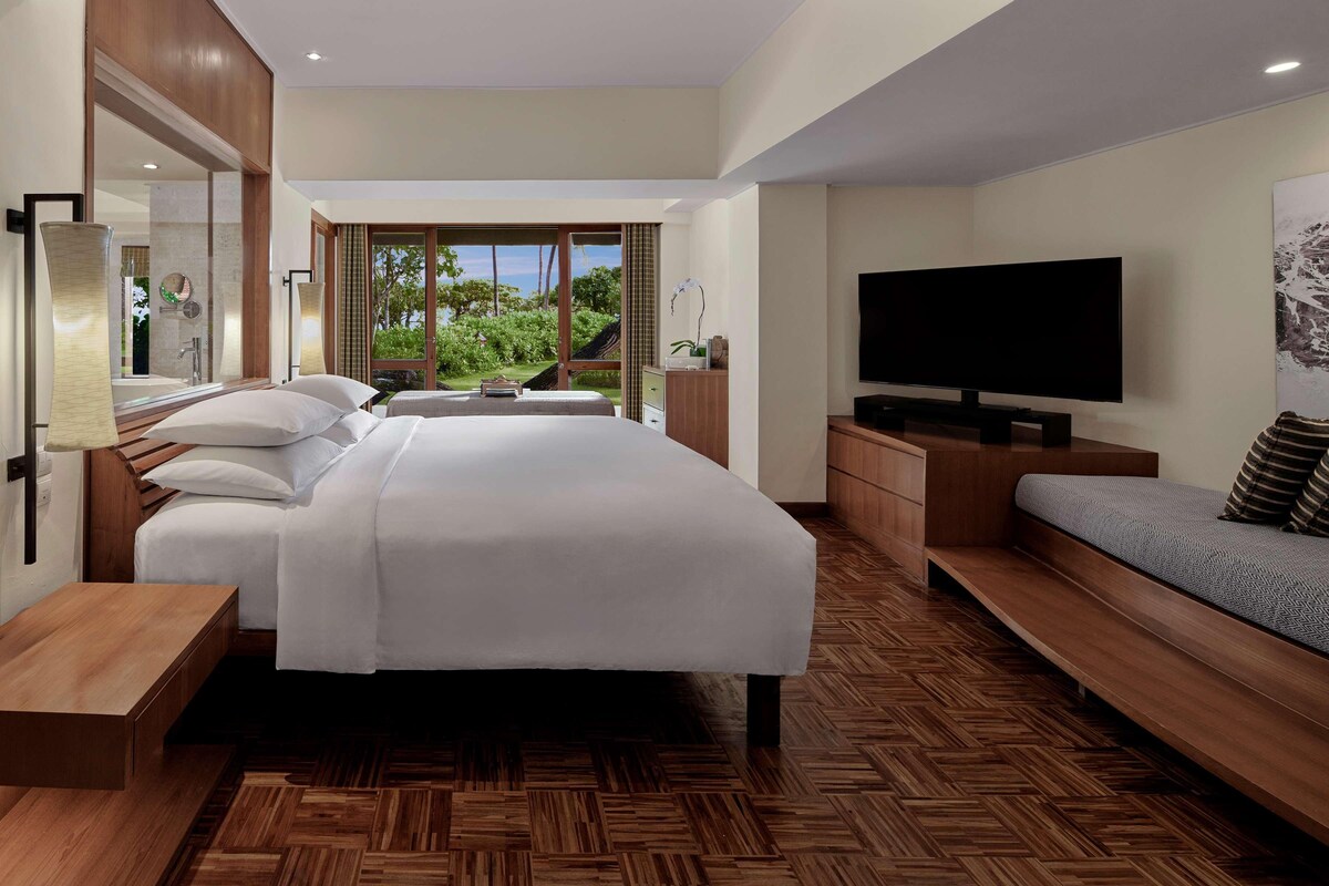 A modern hotel room with a large bed, a TV, a sofa, and a view of greenery outside.