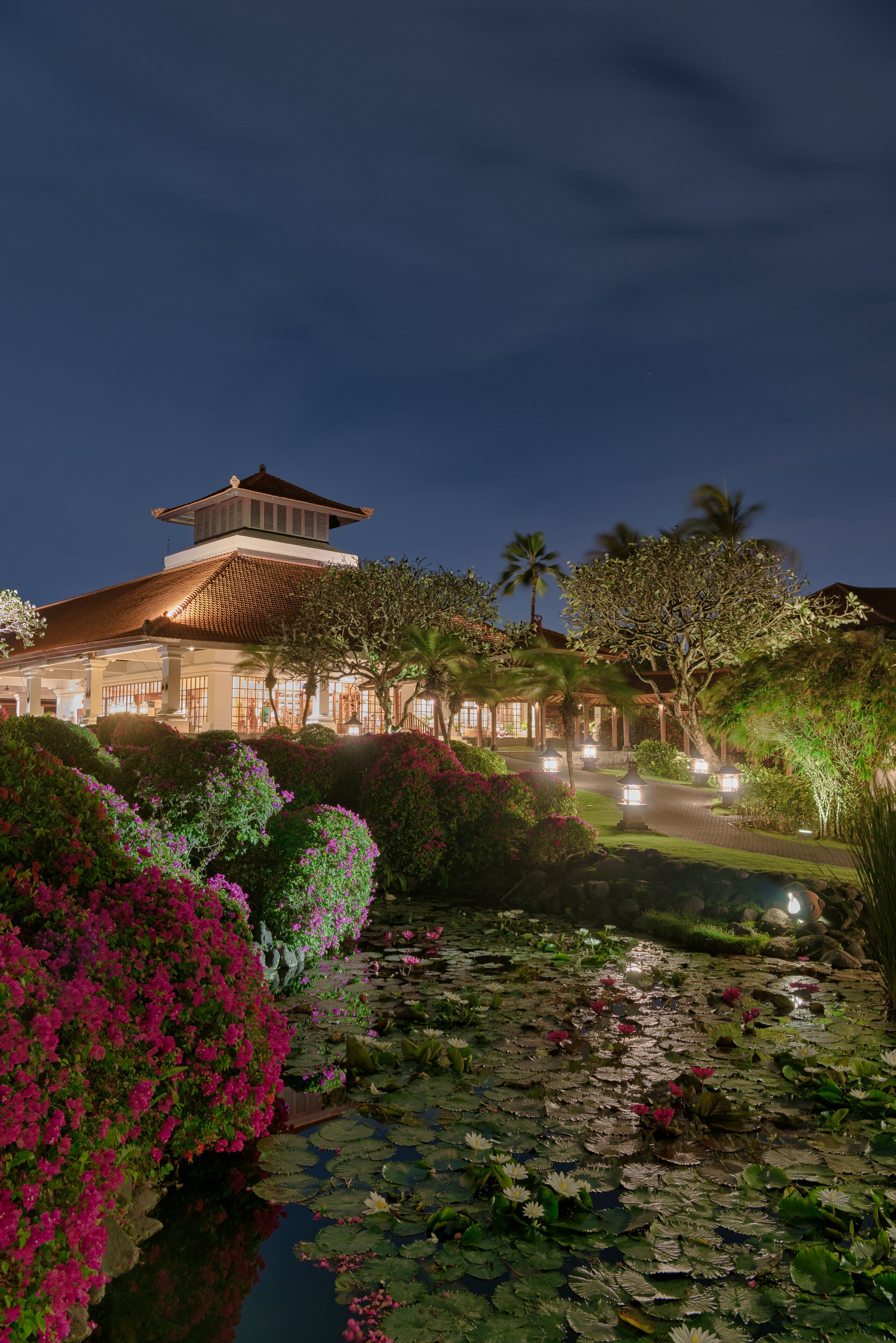 Un edificio tradicional con tejado de tejas y una torre central, rodeado de jardines exuberantes y un estanque con nenúfares.