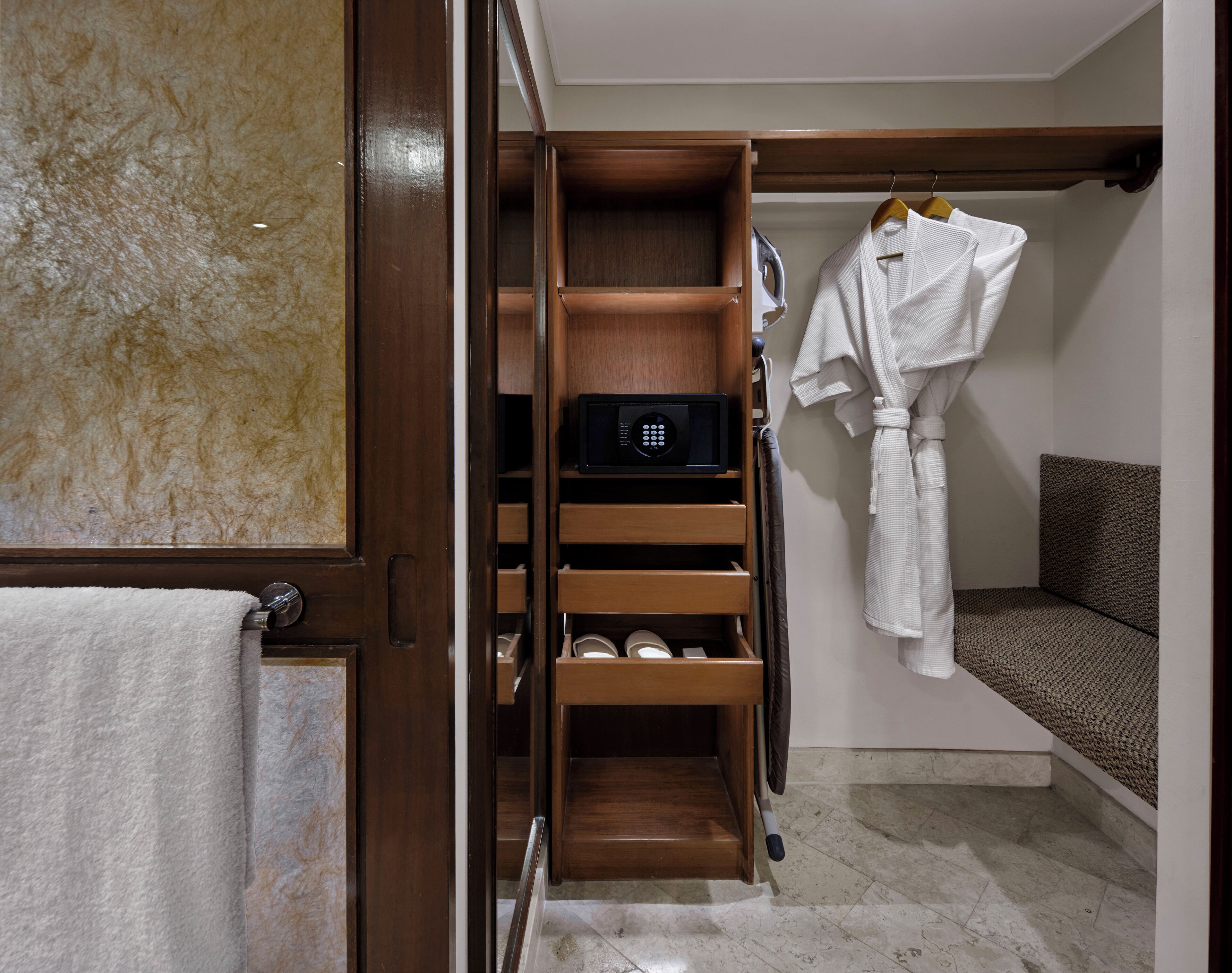 A hotel room with a wooden wardrobe, a robe hanging on the wall, and a small seating area.