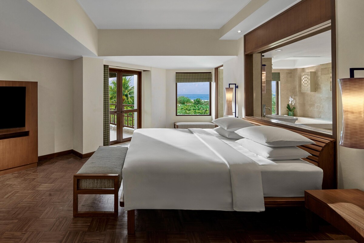 A hotel room with a large bed, a television, a bench, and a view of the ocean.