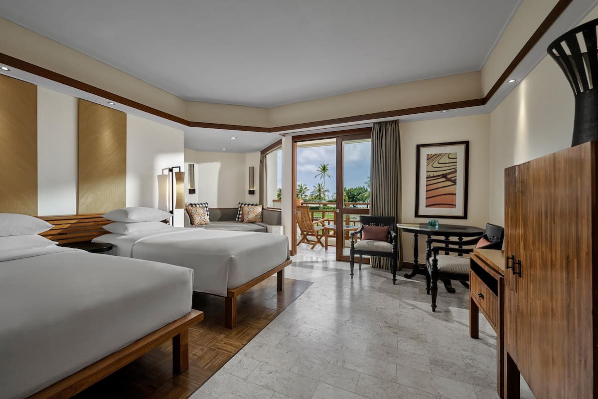 A hotel room with two beds, a desk, and a chair. There is a balcony with a view of palm trees.