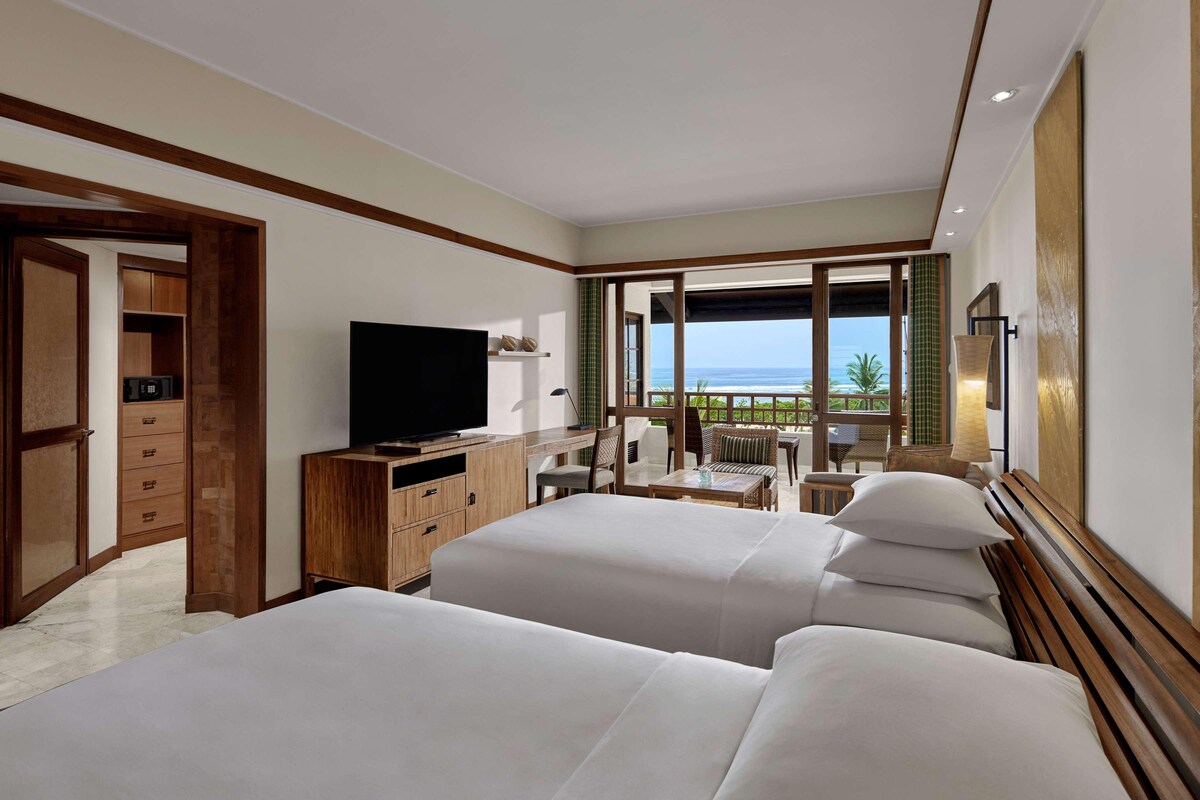 A hotel room with two beds, a TV, a desk, and a balcony with a view of the ocean.