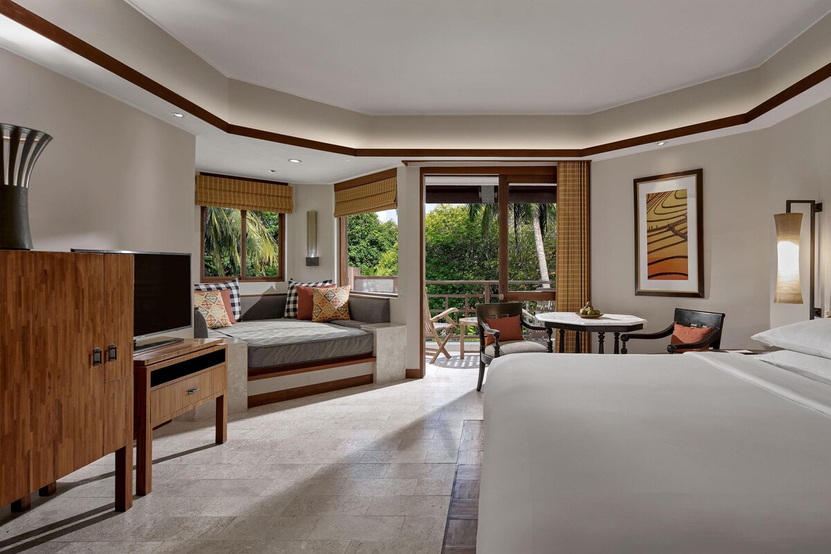 A hotel room with a large bed, a TV, a desk, and a balcony with a view of greenery.