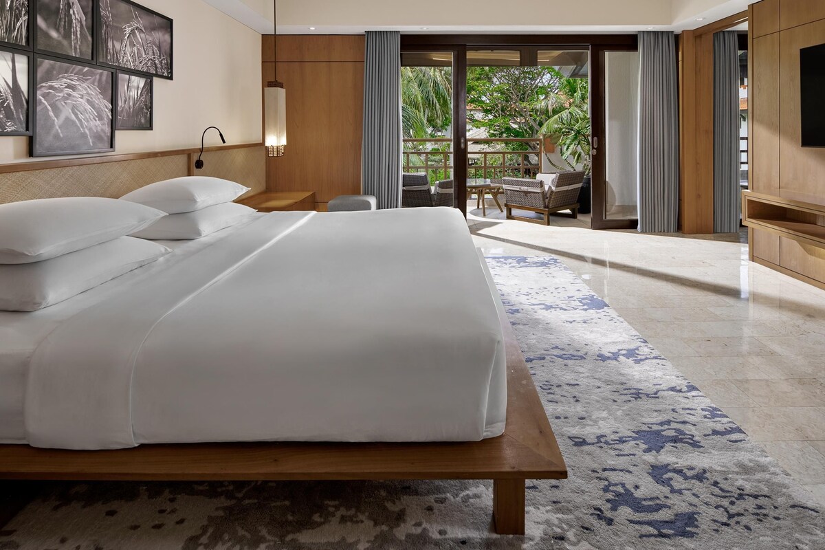 A hotel room with a large bed, a wooden headboard, a flat-screen TV, and a view of a balcony with outdoor furniture.
