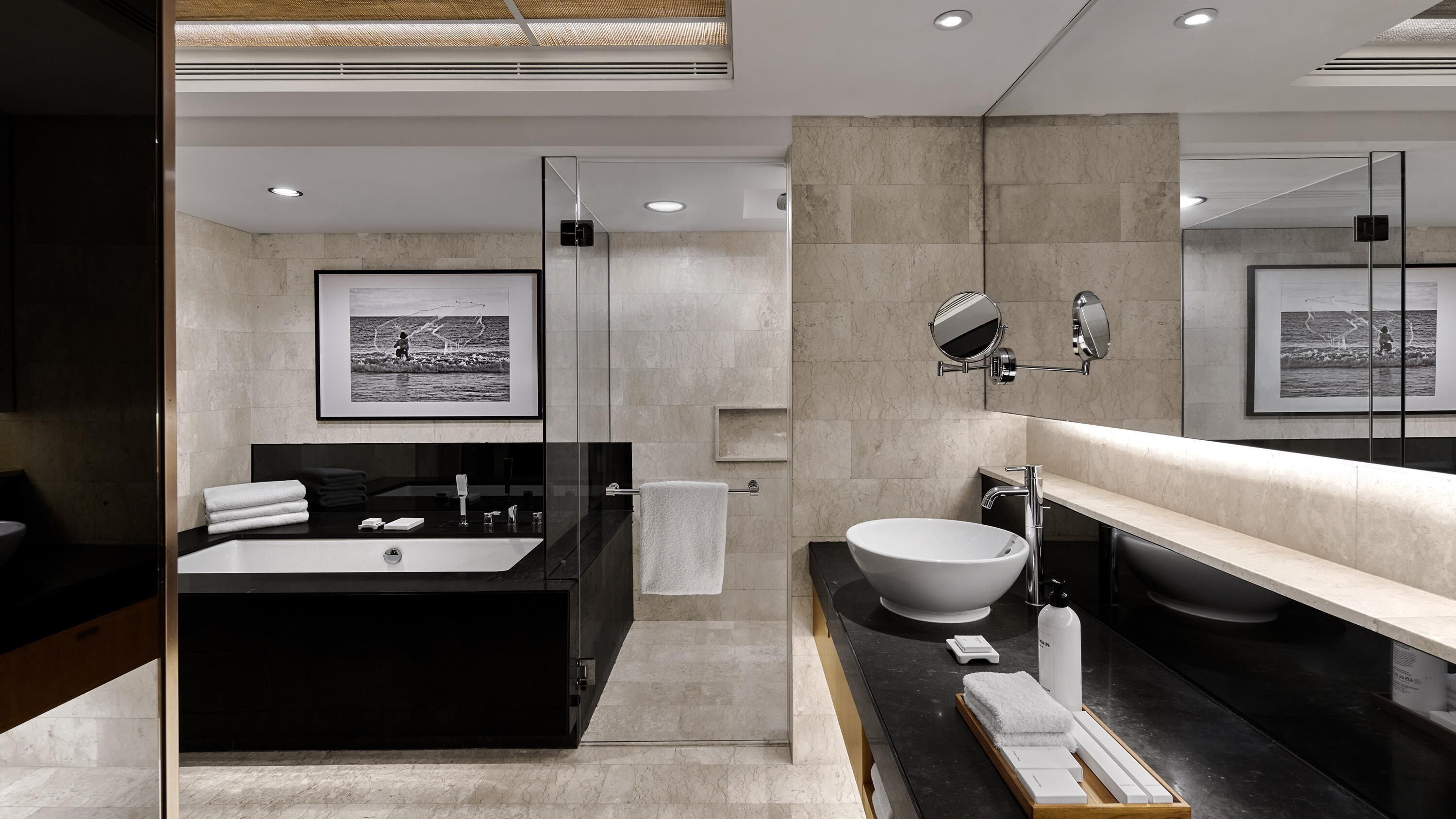 A modern bathroom with a large black bathtub, a white sink, and a large mirror. There are framed pictures on the wall and a towel rack.