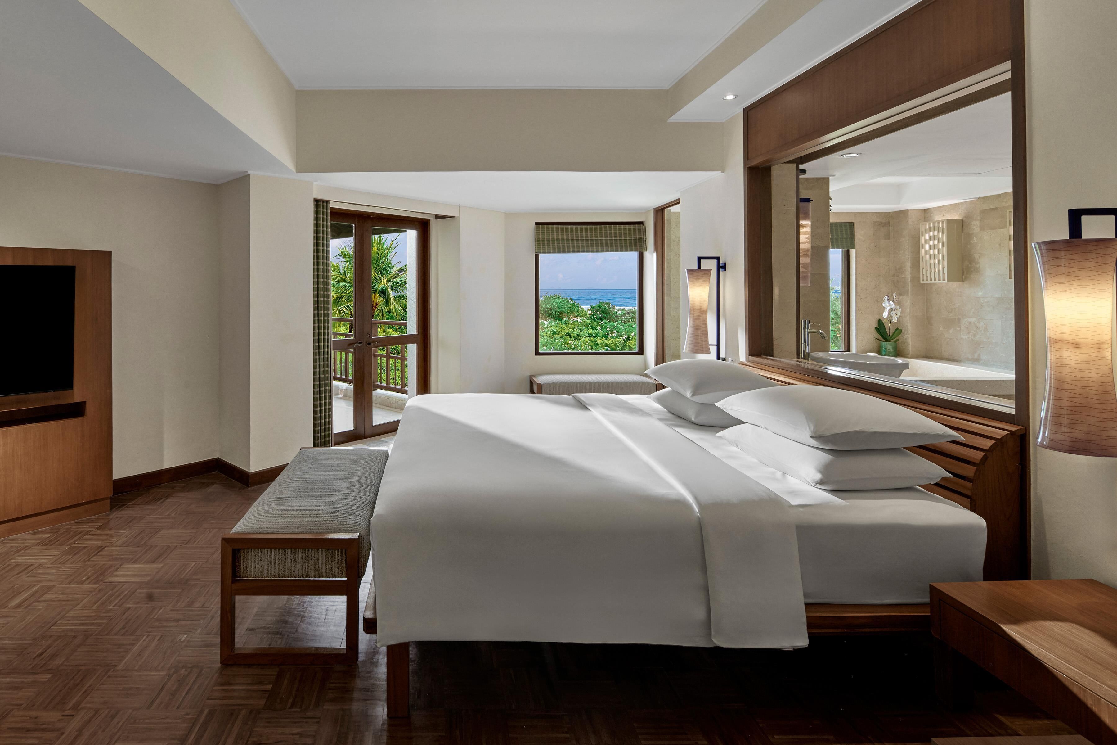 A hotel room with a large bed, a TV, a wooden bench, and a balcony with a view of the ocean.