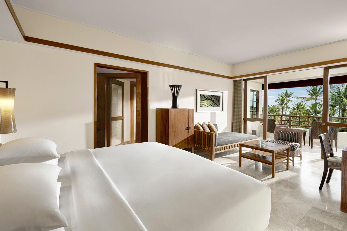 A hotel room with a large bed, a wooden wardrobe, a seating area with a sofa and chairs, and a view of palm trees through the glass door.