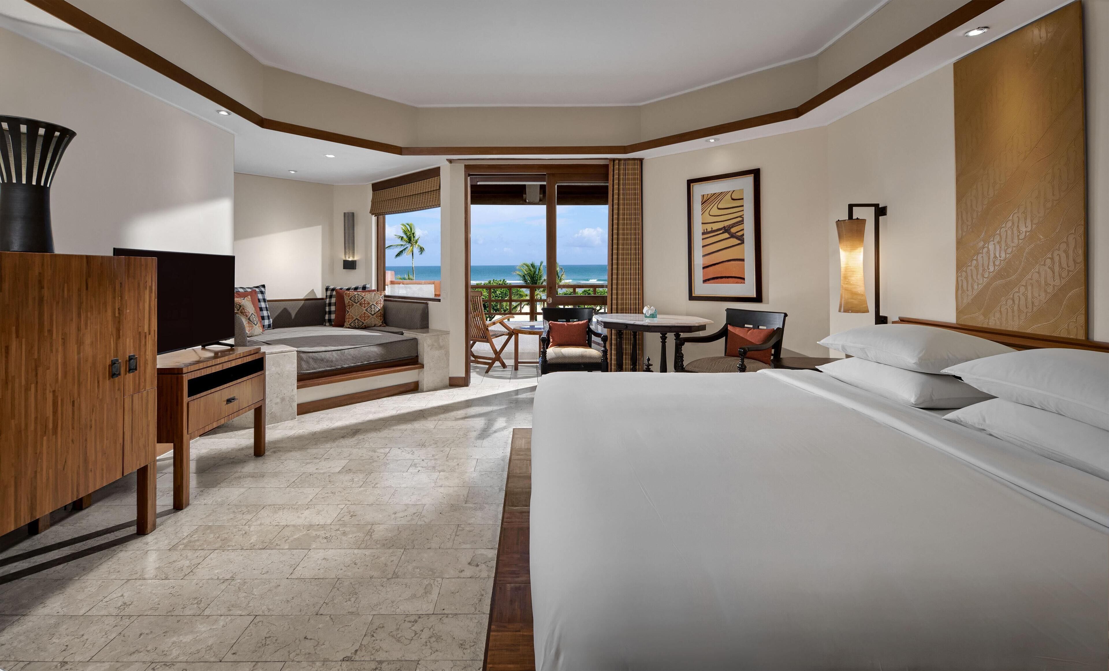 Habitación de hotel con una cama grande, un escritorio con televisor, un área de descanso con vista al océano y un balcón con vista a palmeras.