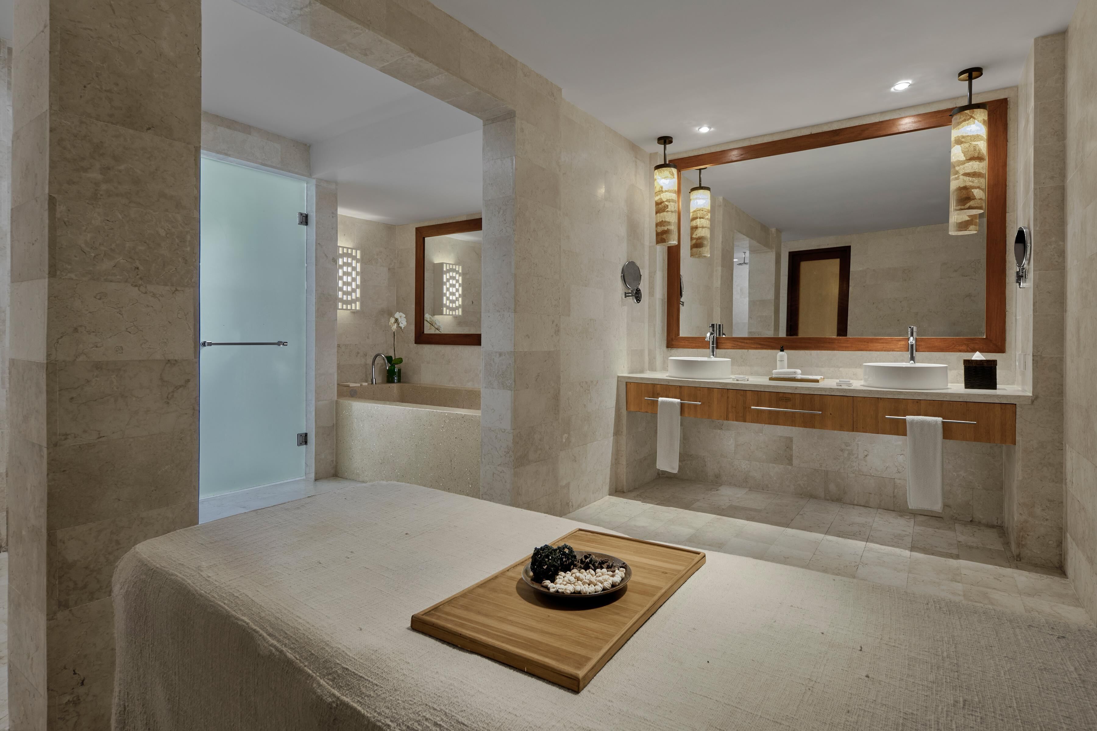 A modern bathroom with a large mirror, two sinks, and a wooden tray with decorative items on the floor.