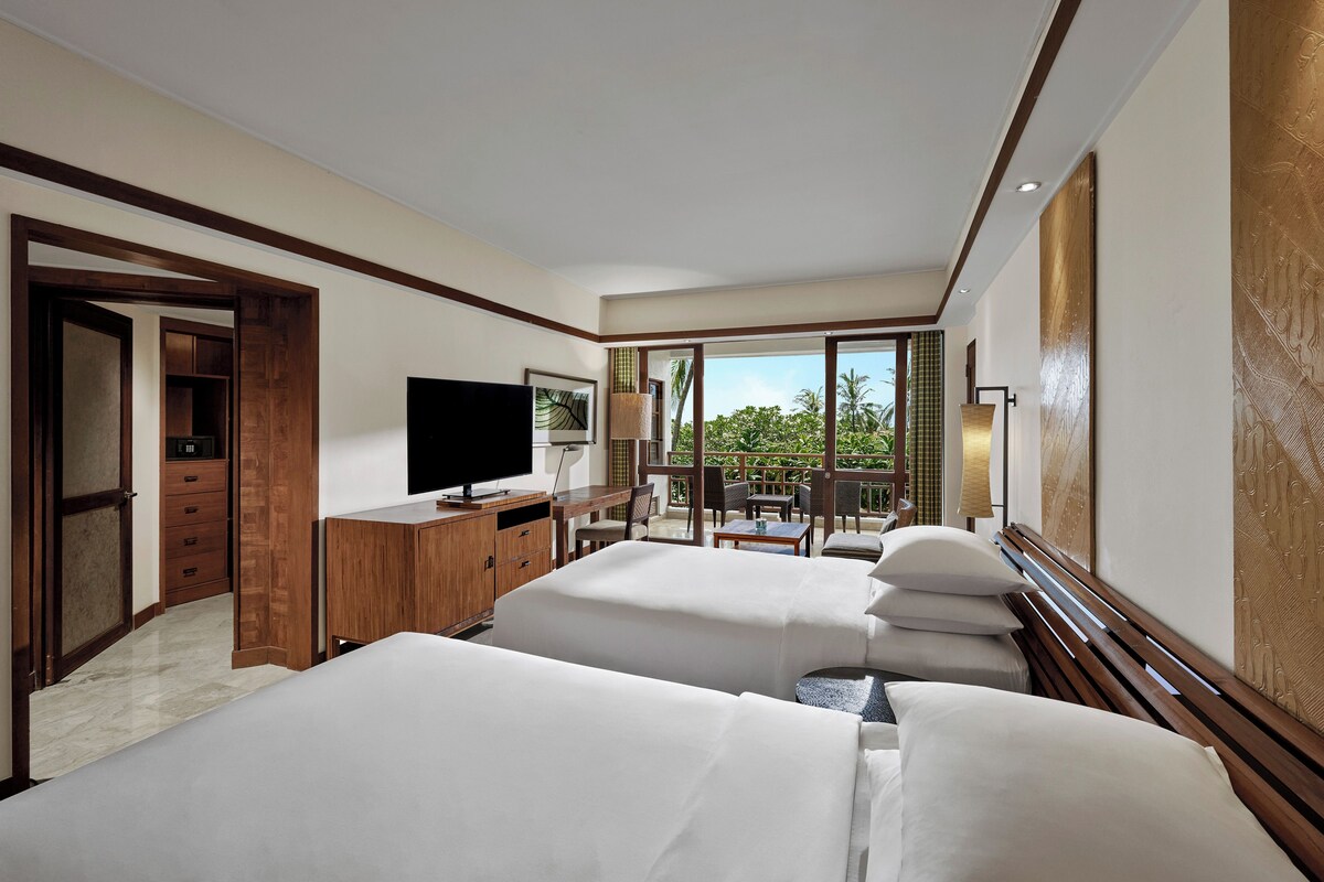 A hotel room with two beds, a TV, a desk, and a balcony with a view of greenery.