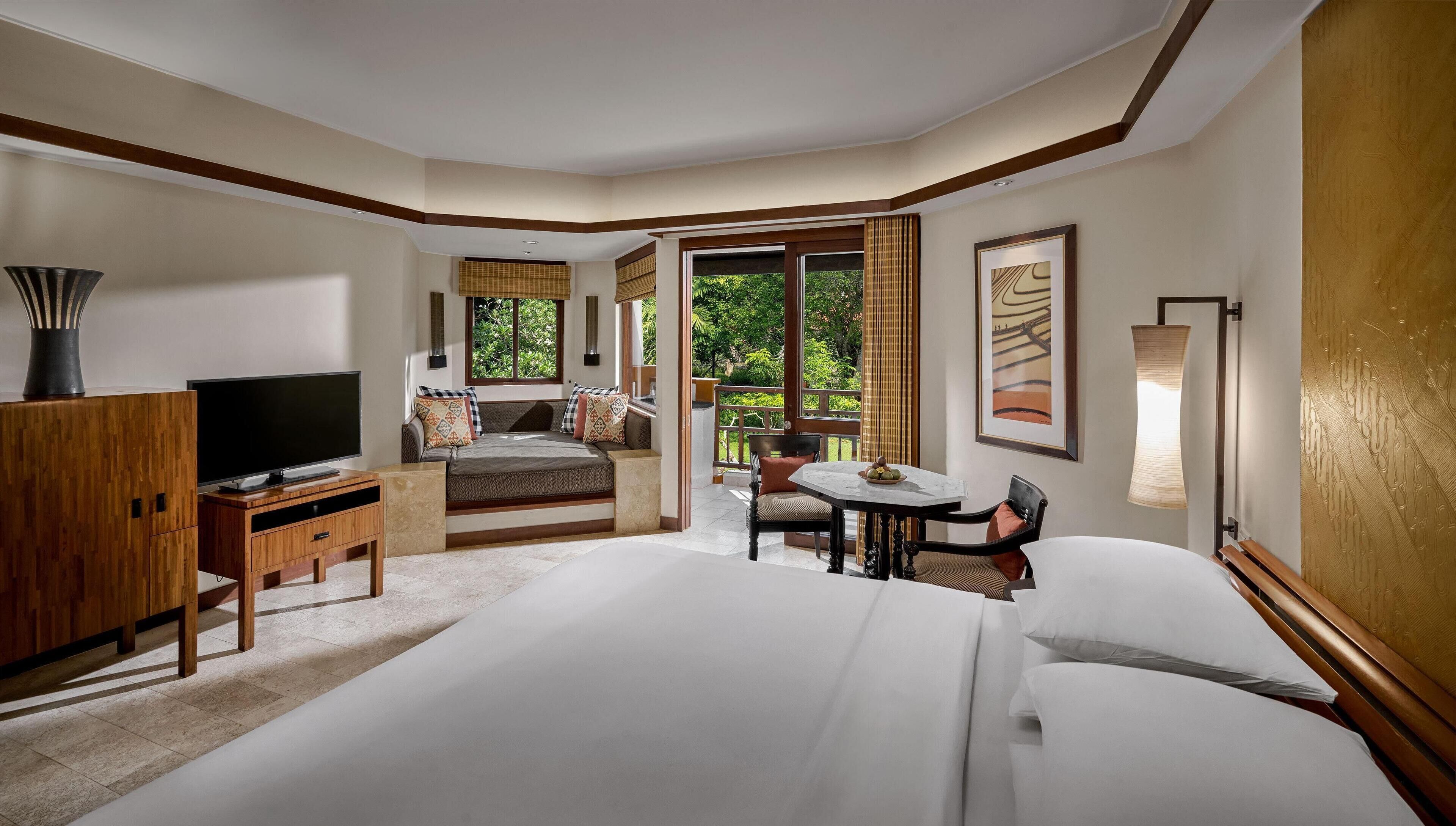 A hotel room with a large bed, a TV, a desk, and a balcony with a view of greenery.