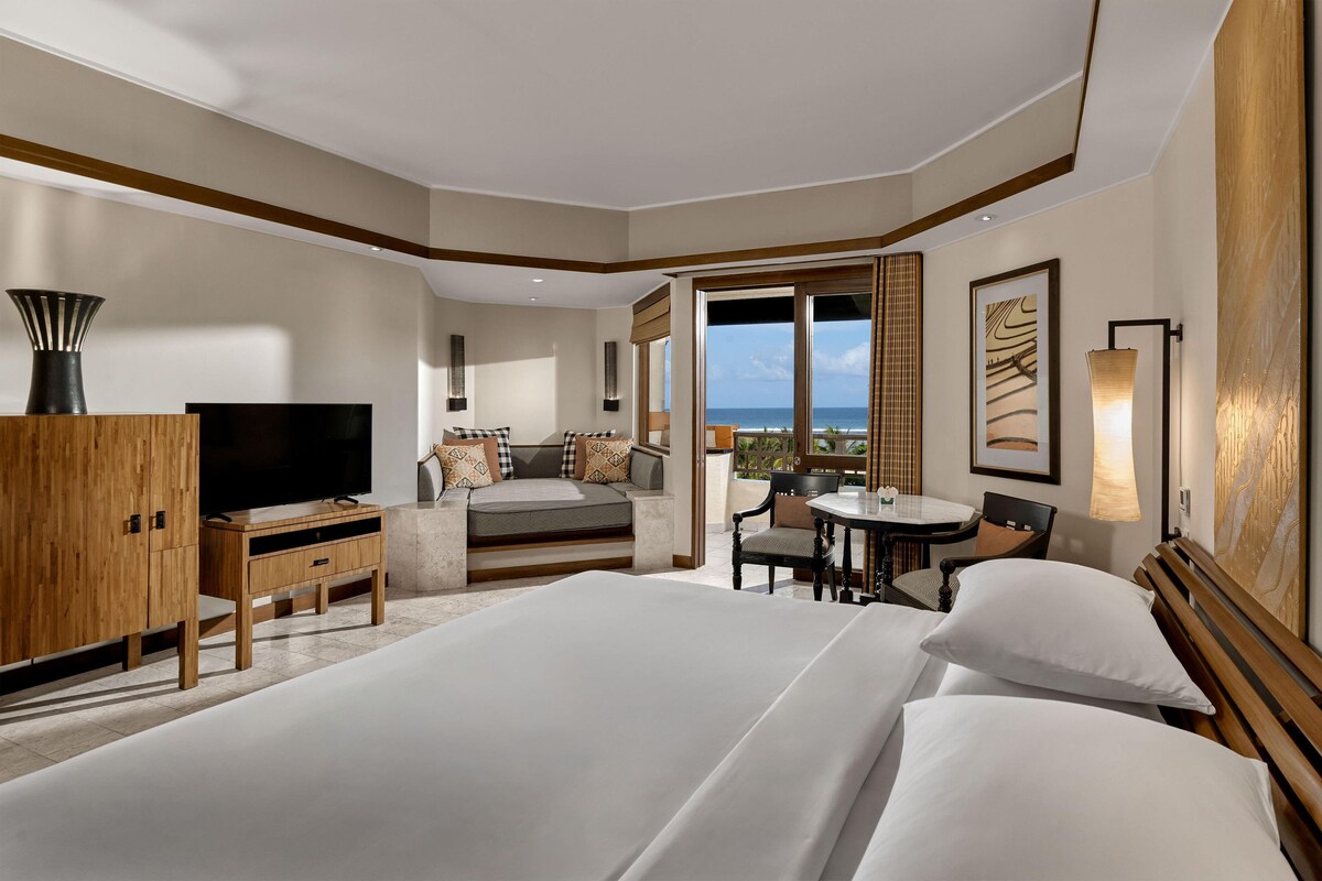 A hotel room with a large bed, a desk, a chair, a TV, and a view of the ocean.