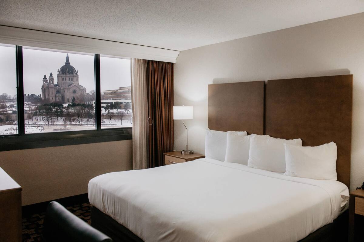 A hotel room with a large bed, a nightstand, a lamp, and a view of a cathedral through the window.