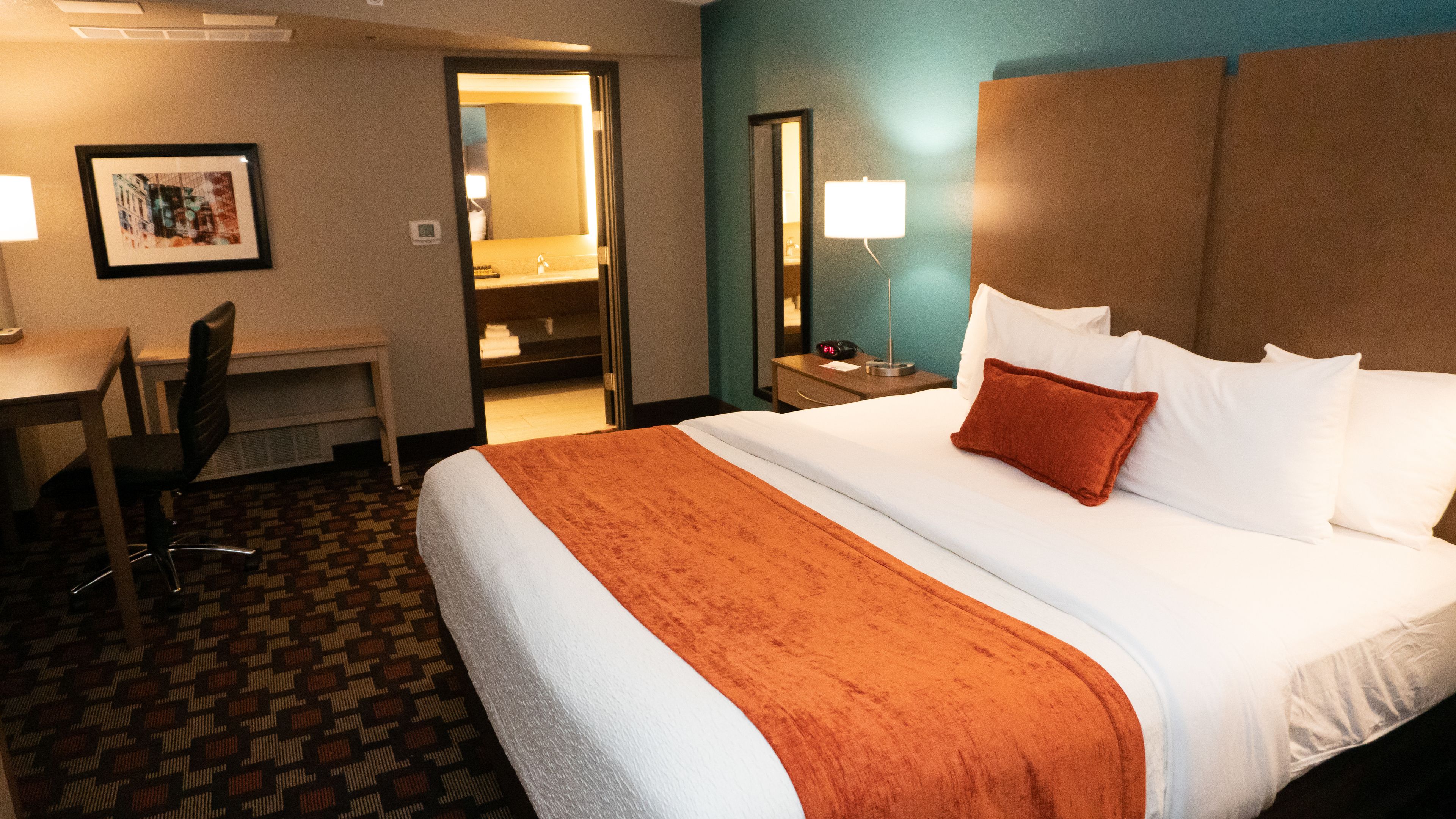A hotel room with a bed, a desk, a chair, a nightstand, and a mirror.