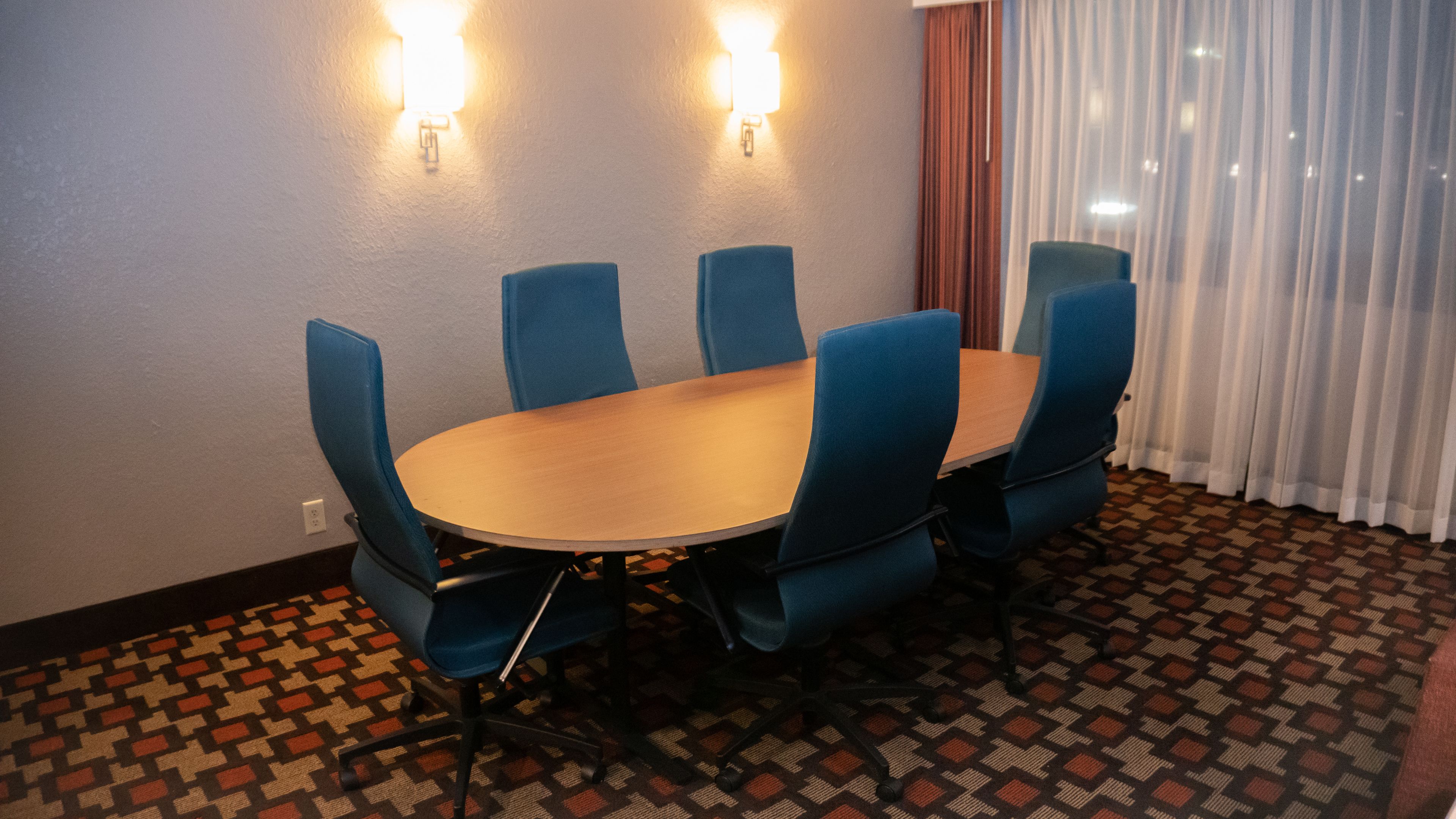 Une salle de conférence avec une table ronde et six chaises bleues, deux lampes fixées au mur et un tapis à motifs.