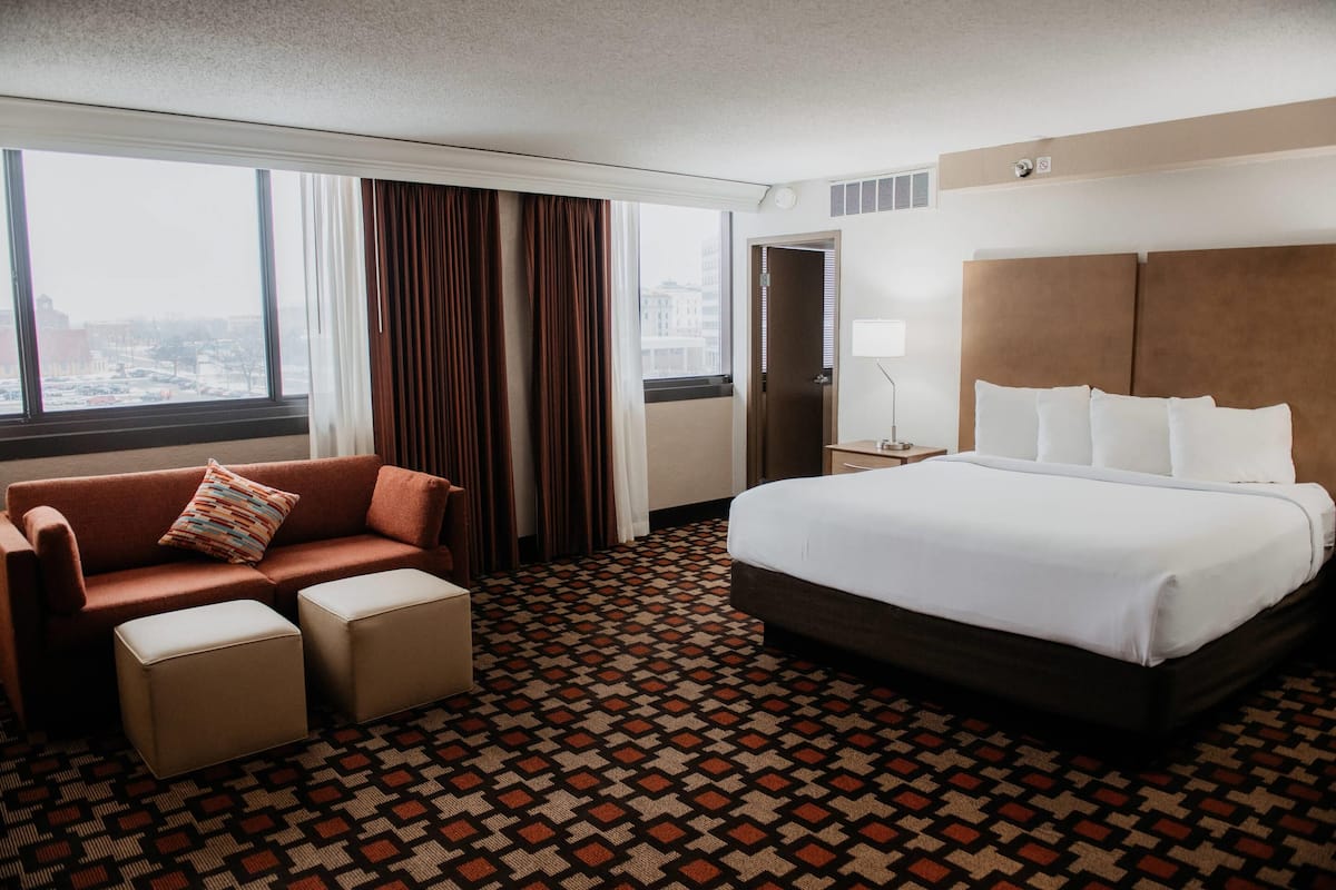 A hotel room with a large bed, a sofa, two ottomans, a nightstand, and a view of the city through large windows.