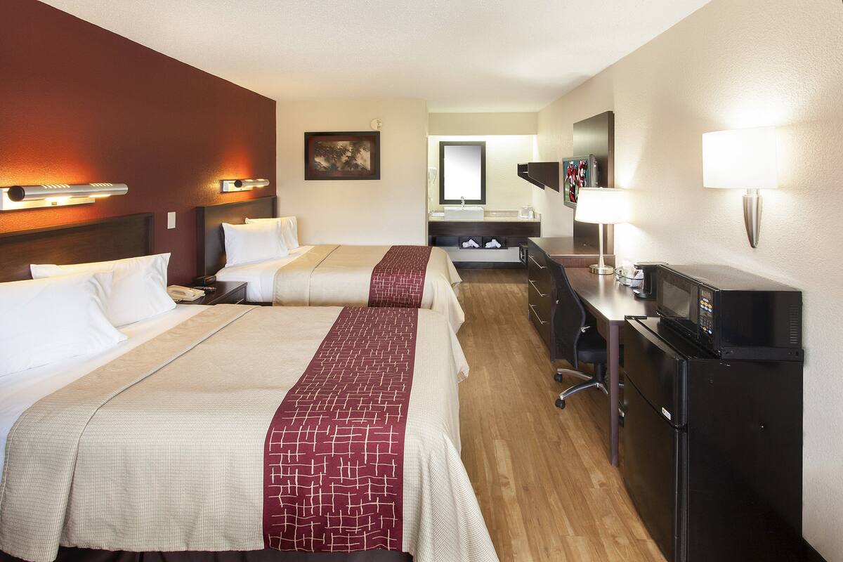 A hotel room with two beds, a desk, a refrigerator, and a bathroom in the background.