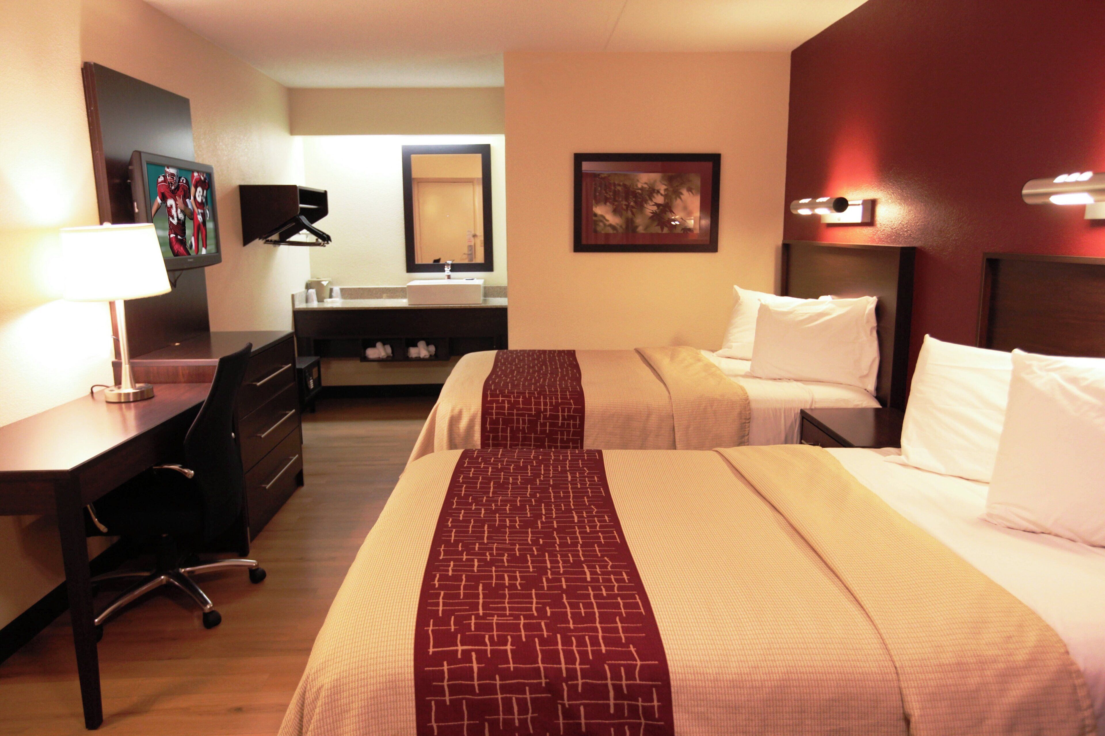 Une chambre d’hôtel avec deux lits, un bureau, une chaise, une commode, une salle de bain avec un lavabo et un miroir, et un tableau accroché au mur.