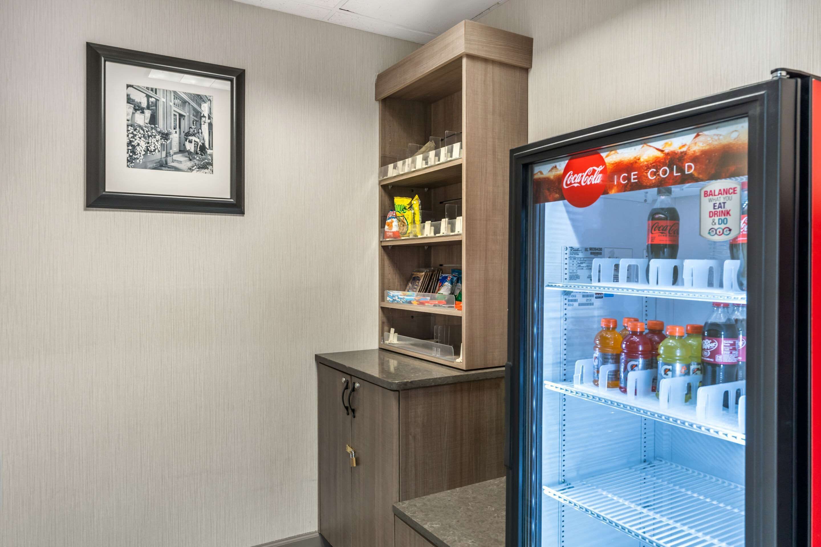 A hotel room with a refrigerator stocked with beverages, a wooden cabinet with shelves, and a framed black and white photograph on the wall.