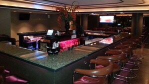 Una zona de bar con una barra alargada, taburetes, una pantalla de televisión y una planta decorativa.