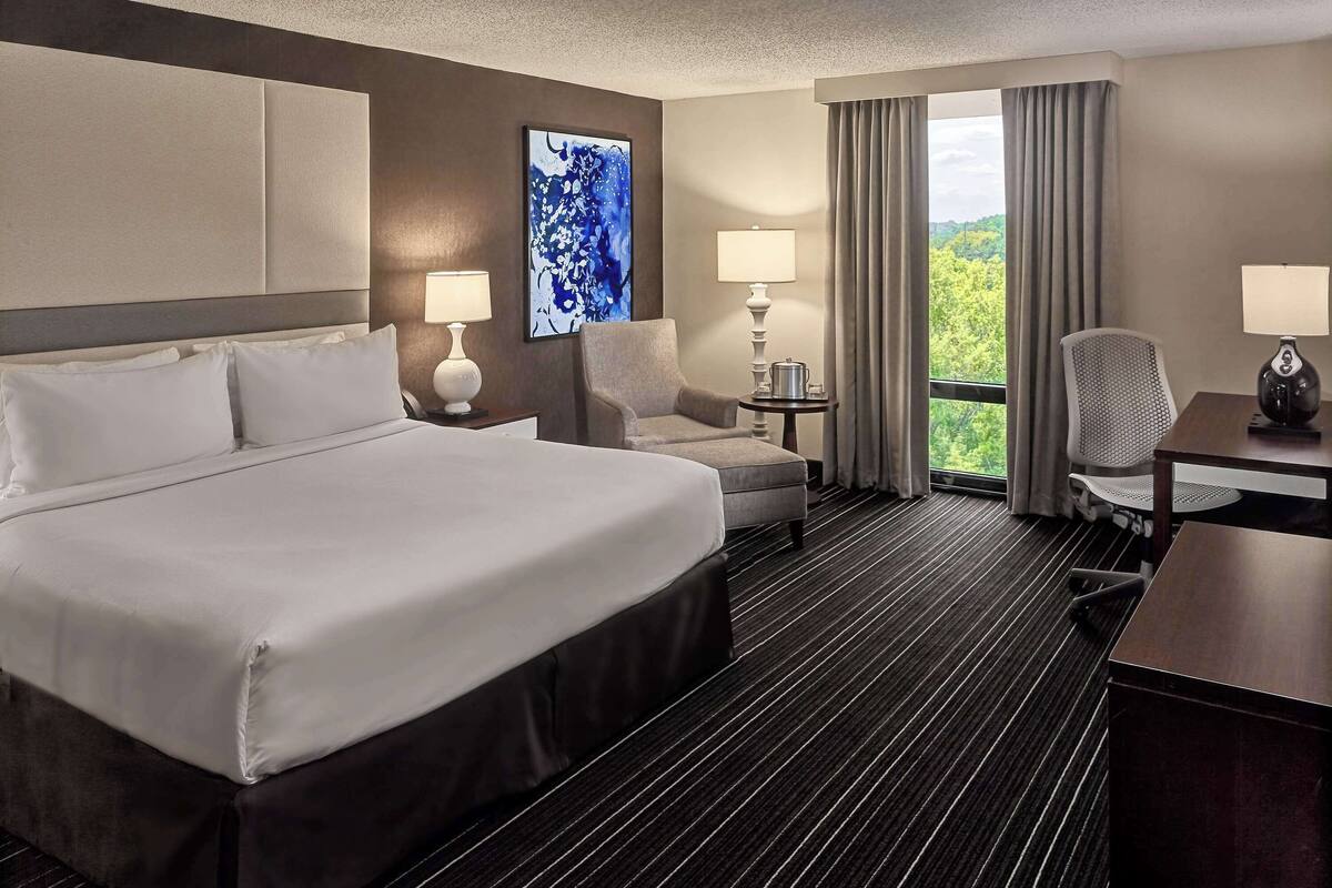 A hotel room with a large bed, a desk, two chairs, a small table with a lamp, and a view of greenery through the window.