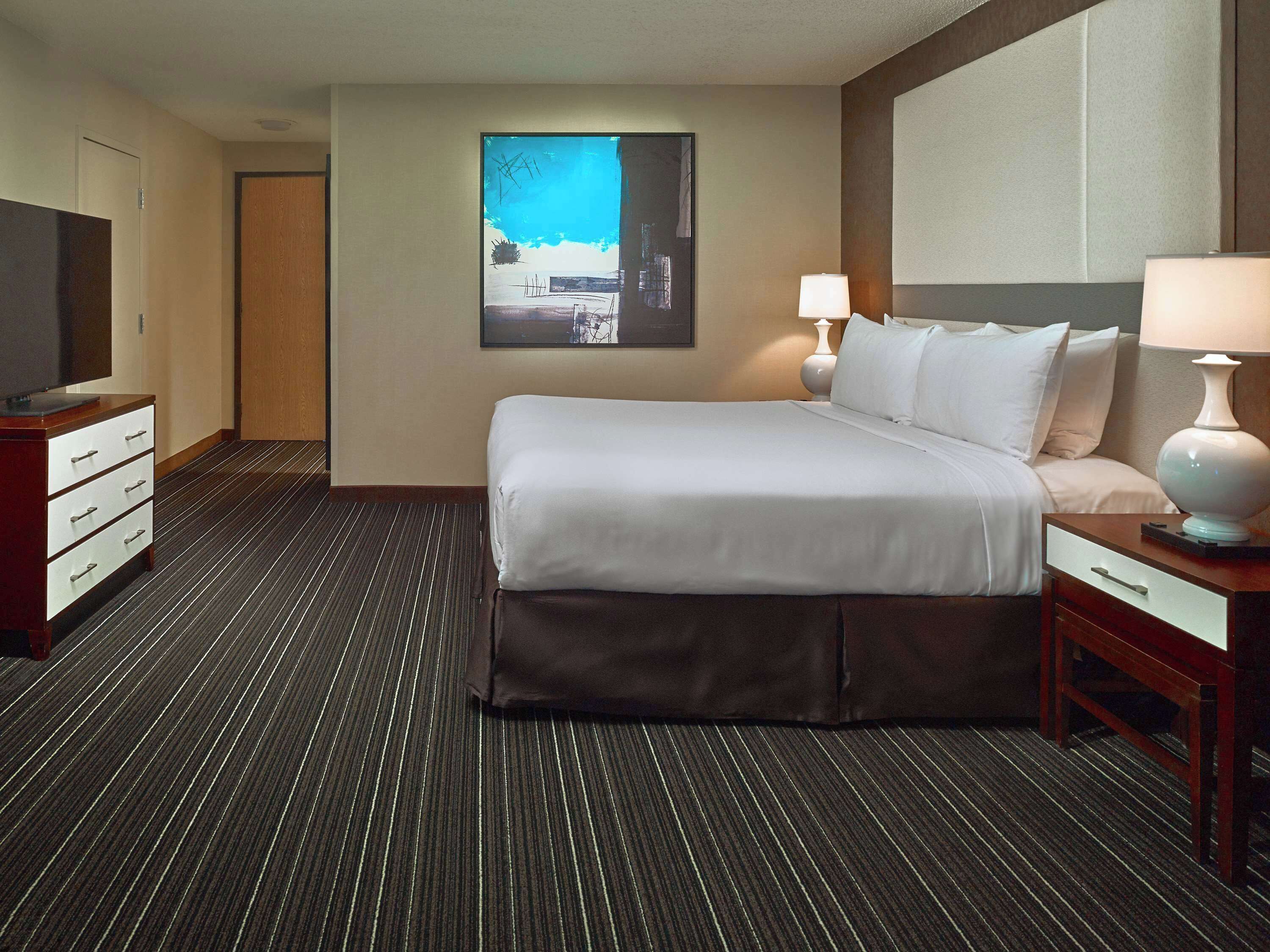 Una camera d'albergo con un letto grande, due comodini con lampade, una TV a schermo piatto su un comò e un quadro appeso al muro.
