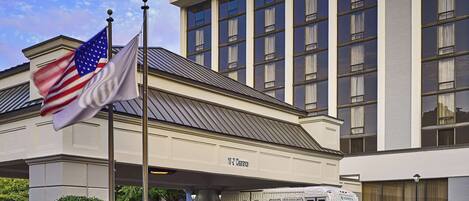 A hotel entrance with a flagpole, a parked shuttle bus, and a clear sky.
