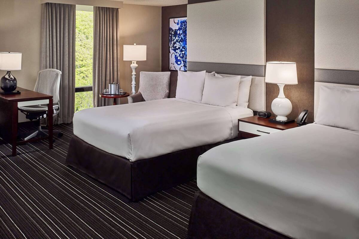 A hotel room with two beds, a desk, a chair, a lamp, and a view of greenery.