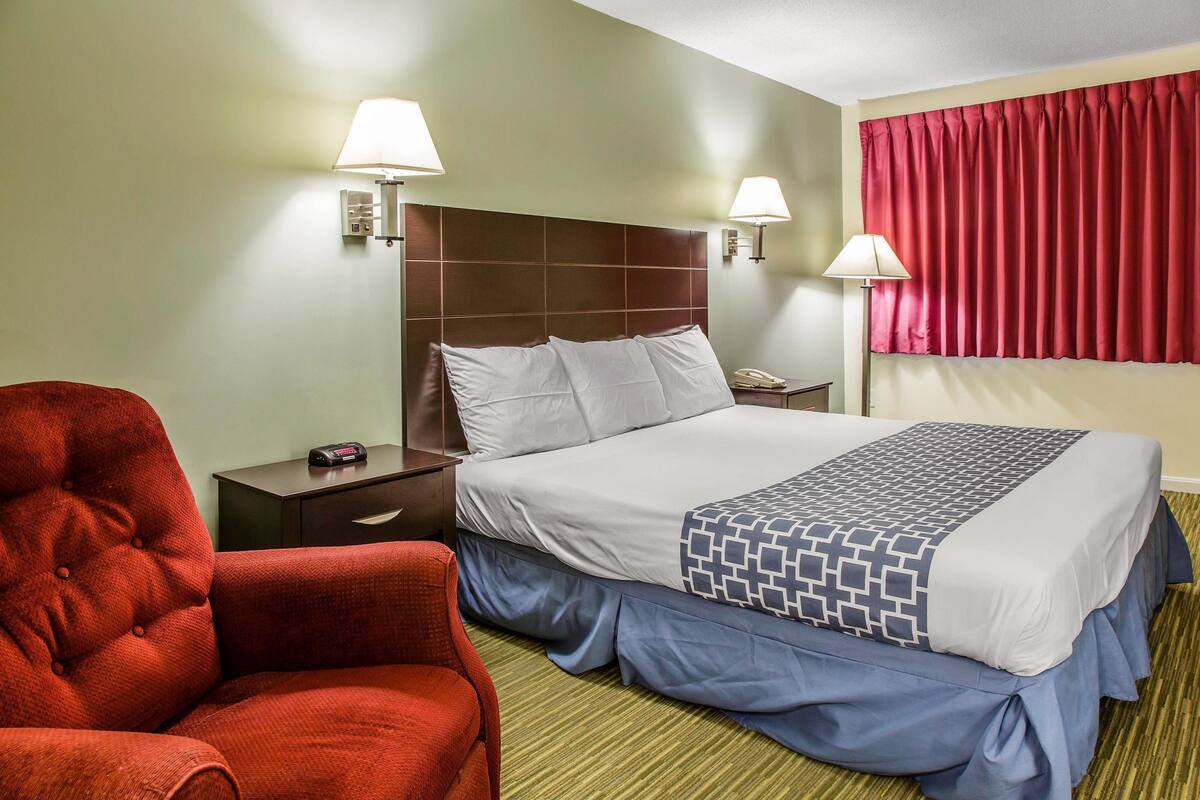 Una habitación de hotel con una cama grande, dos lámparas de noche, una silla roja y una mesita de noche con un teléfono y un control remoto.
