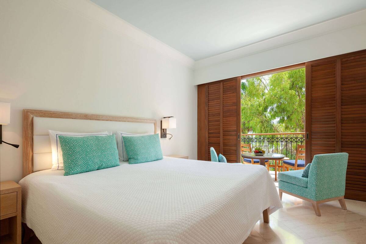 Habitación de hotel con una cama grande, cabecera de madera, balcón con mesa y sillas, y vista a zonas verdes.