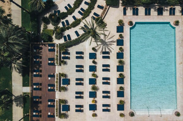 An aerial view of a resort with a large swimming pool, multiple lounge areas, and palm trees.