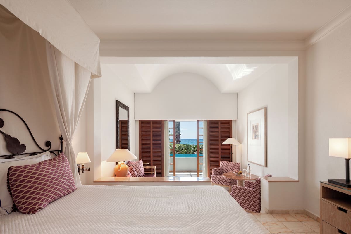 Una habitación de hotel con una cama grande, una zona de estar con una mesa y sillas, un balcón con vistas al océano y un espejo grande en la pared.