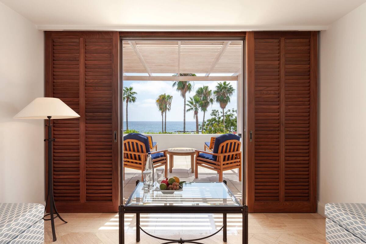 Una habitación con una mesa de vidrio, dos sillas, una lámpara y vistas a palmeras y al mar.