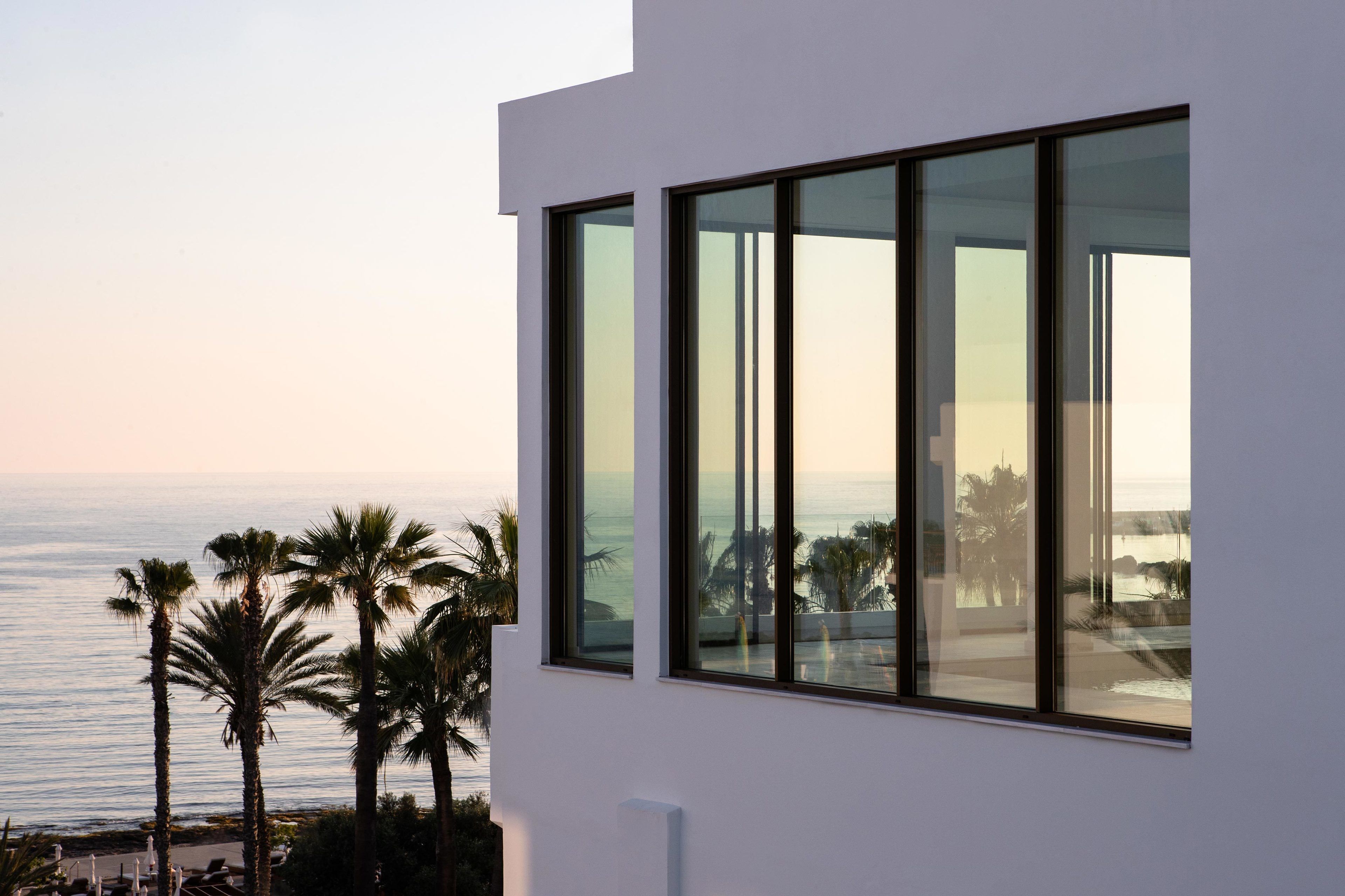 Un edificio moderno con grandes ventanales que dan a una playa con palmeras.