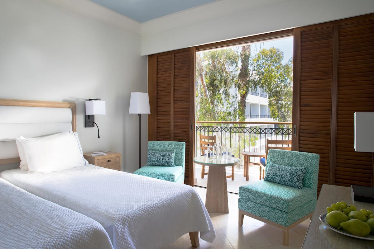 Habitación de hotel con dos camas, un balcón con contraventanas de madera, una mesa redonda, dos sillas y un frutero con fruta.
