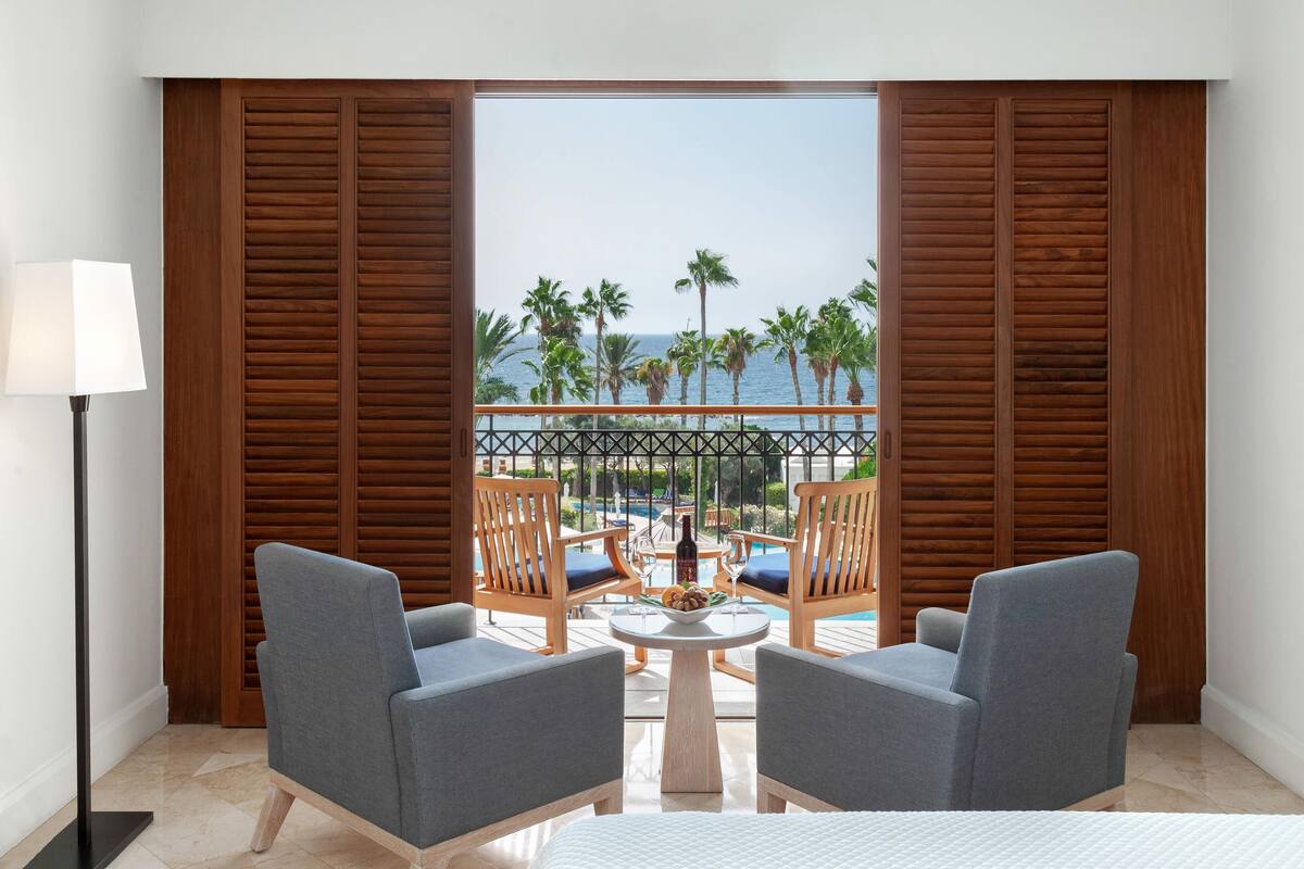 Una habitación con dos sillones grises, una mesita redonda con un frutero, y vistas al océano y palmeras a través de contraventanas de madera.