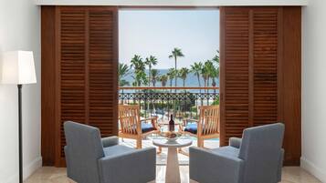 Ein Zimmer mit zwei grauen Sesseln, einem kleinen runden Tisch mit einer Obstschale und Blick auf das Meer und Palmen durch Holzjalousien.