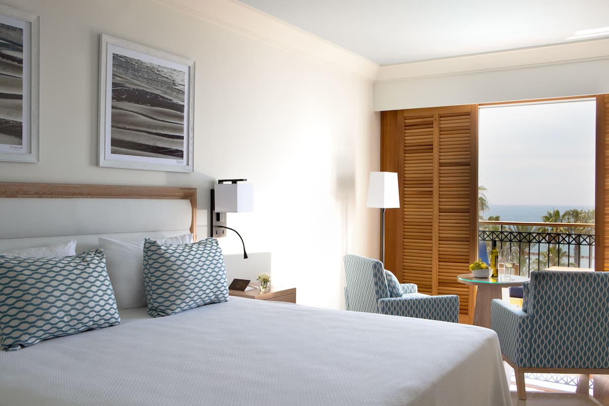 Habitación de hotel con una cama grande, dos almohadas estampadas, una mesita de noche con lámpara, una pequeña mesa con un florero y una botella, y vista al océano a través de un balcón con persianas de madera.