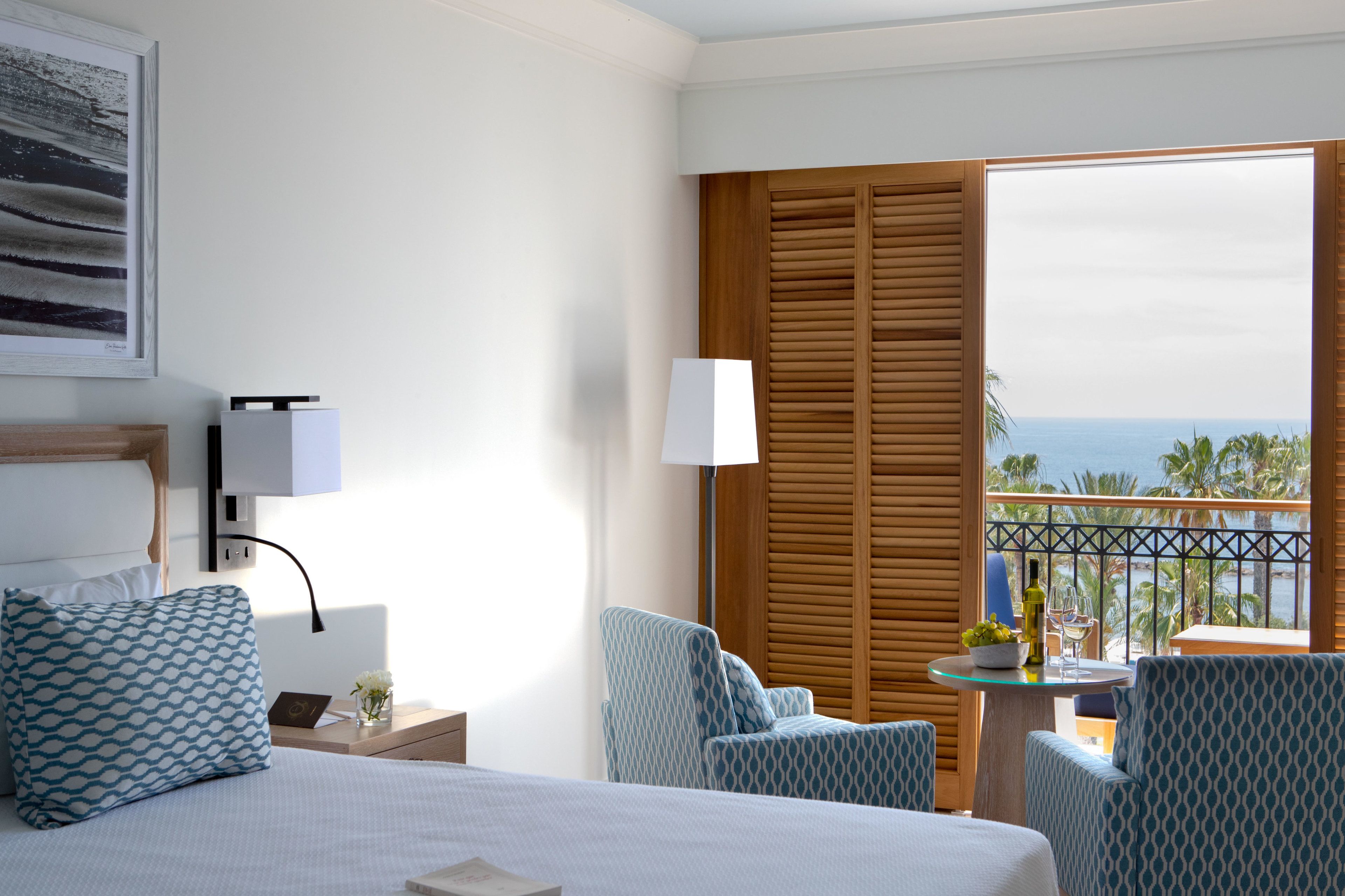 Une chambre d’hôtel avec un lit, deux fauteuils à motifs, une petite table avec une bouteille et un verre, et une vue sur l’océan depuis un balcon doté de volets en bois.