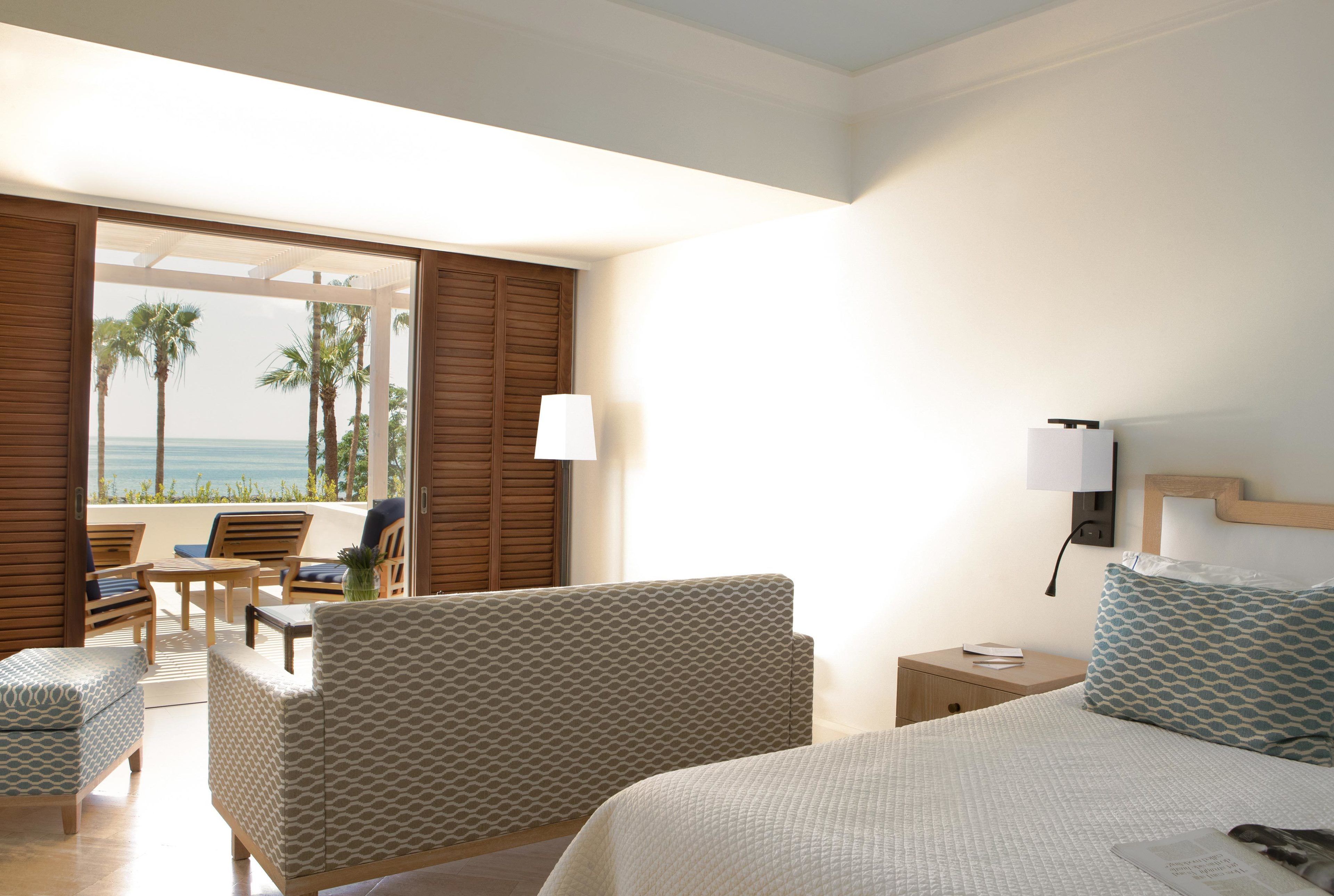 Habitación de hotel con cama, dos sillas, una mesita y una mesita de noche. Se ve una vista de palmeras y el océano a través de una puerta abierta.