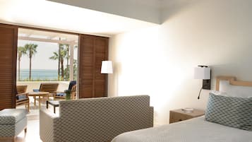 Habitación de hotel con cama, dos sillas, una mesita y una mesita de noche. Se ve una vista de palmeras y el océano a través de una puerta abierta.