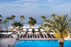 A poolside area with lounge chairs and umbrellas, palm trees, and a view of the sea.