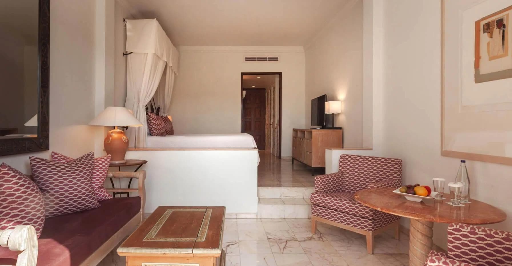 A hotel room with a bed, a sofa, a chair, a small table, a lamp, and a TV.