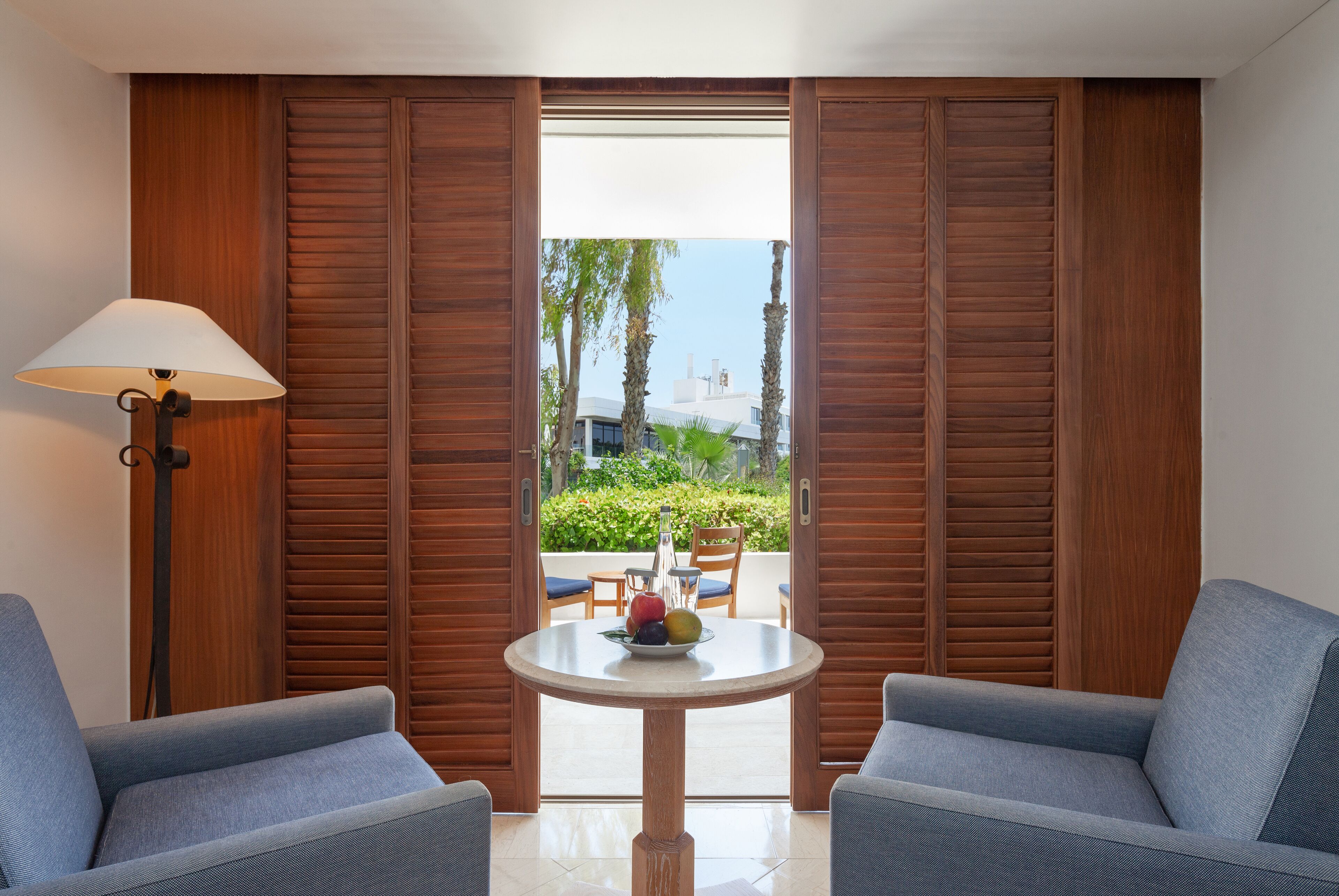 A room with a round table, two blue armchairs, a lamp, and wooden shutters.