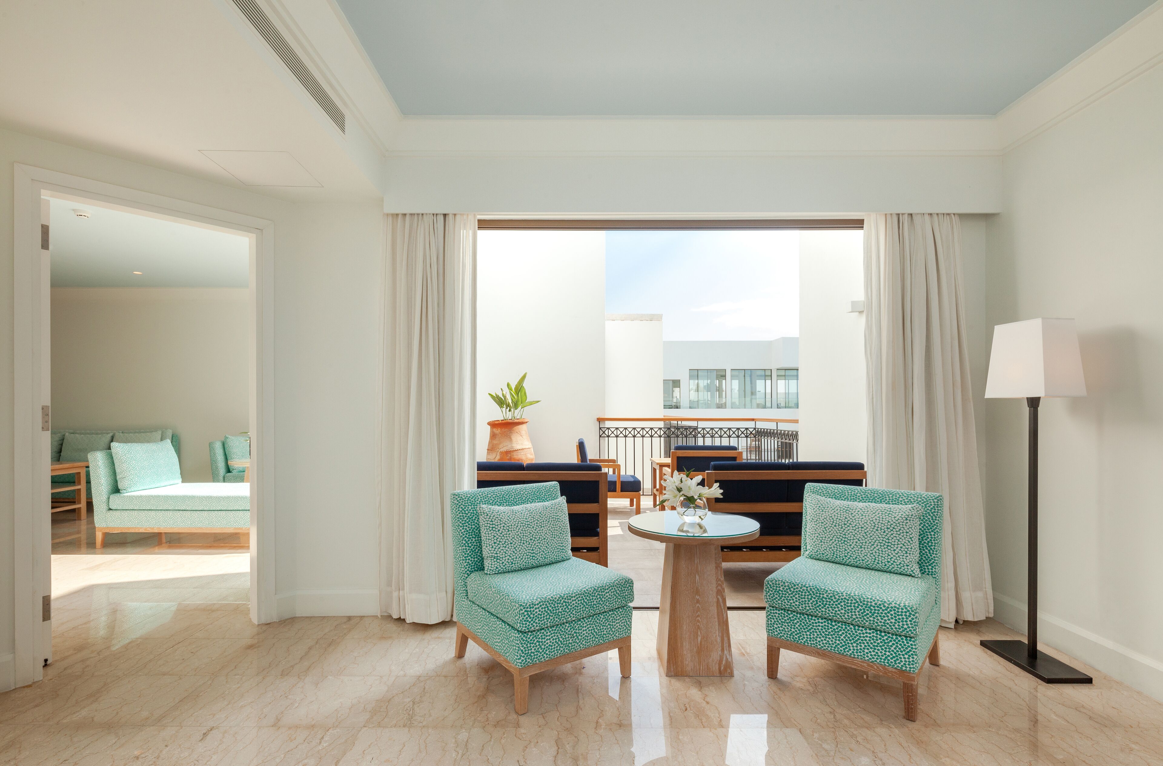 A hotel room with a balcony, two patterned armchairs, a round coffee table, and a floor lamp.