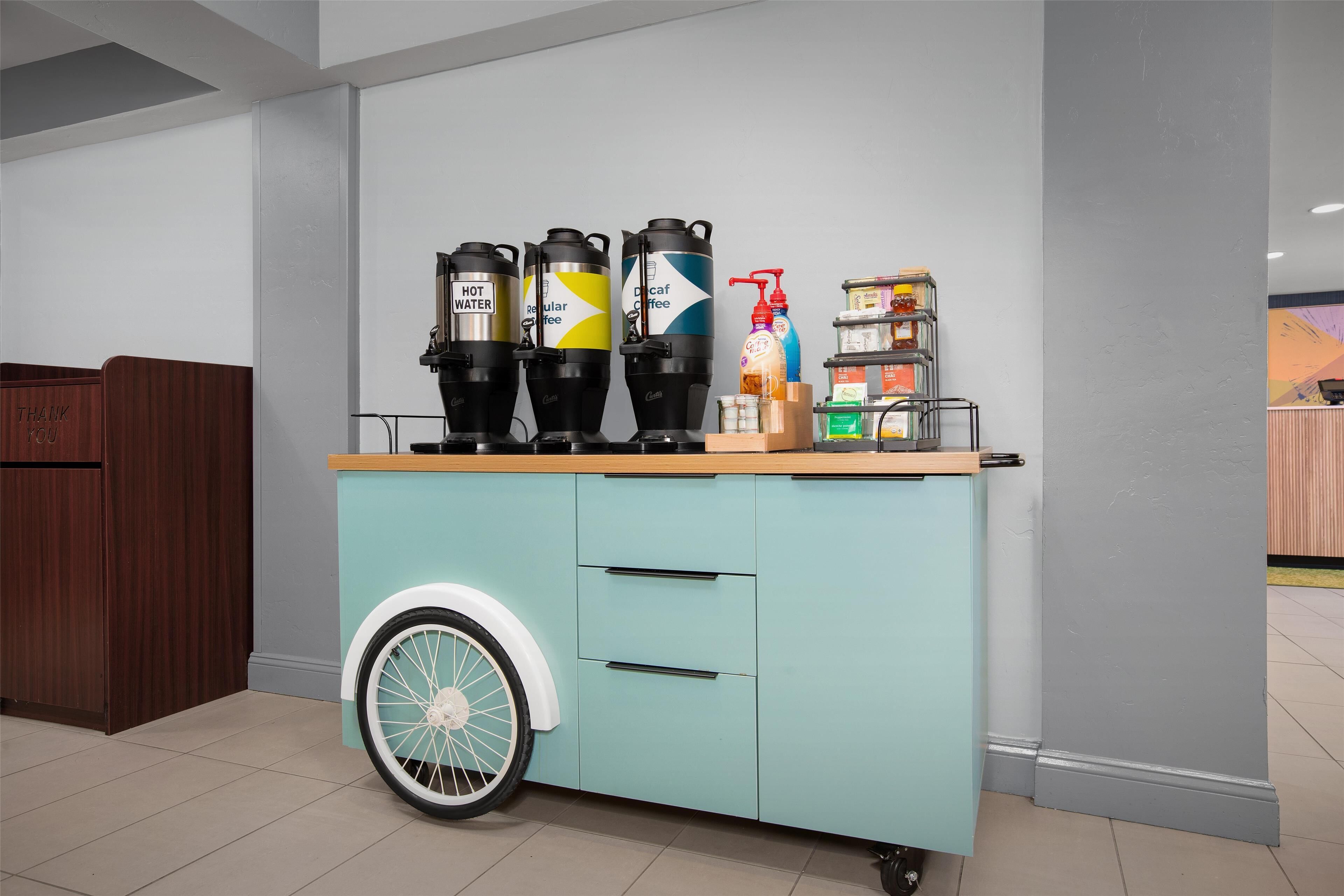 A kitchen area with a teal-colored cabinet featuring a wheel, three coffee dispensers labeled 'HOT WATER,' 'Regular,' and 'Decaf Coffee,' and a bottle of hand sanitizer.