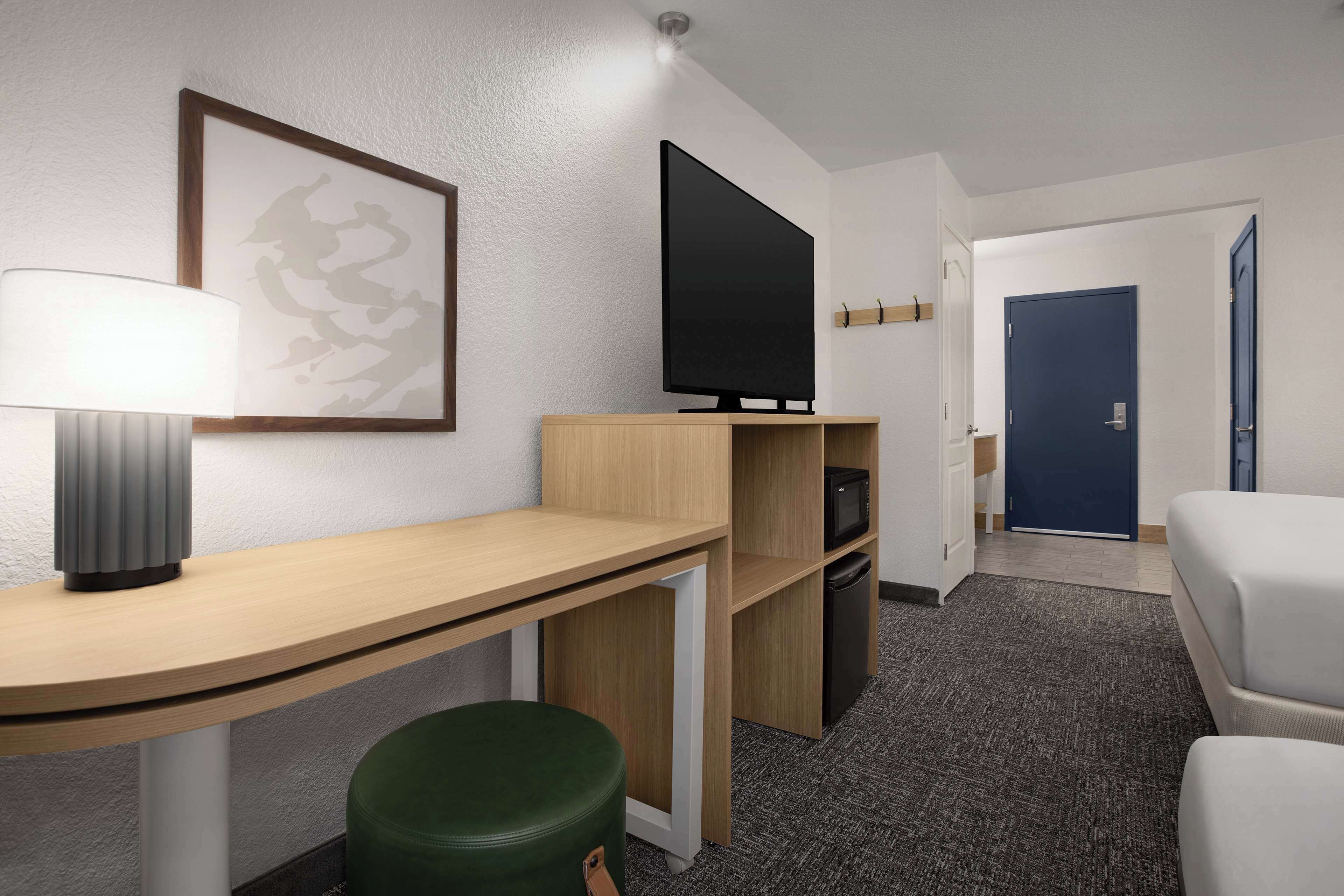 A hotel room with a desk, a green stool, a flat-screen TV, a framed artwork, a white bed, and a blue door.